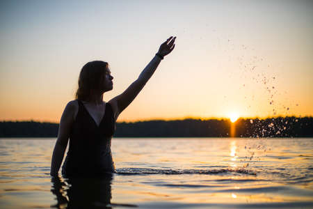 Beautiful Russian girl in a long black swimsuit swims outside the city on the lake in the rays of sunset or dawn. Splashing water on the background of the sun. Happy girl.の写真素材