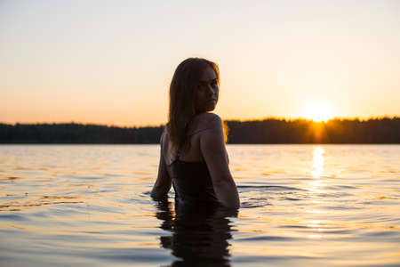 Beautiful Russian girl in a long black swimsuit swims outside the city on the lake in the rays of sunset or dawn. Splashing water on the background of the sun. Happy girl.の写真素材