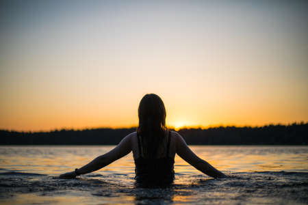 Beautiful Russian girl in a long black swimsuit swims outside the city on the lake in the rays of sunset or dawn. Splashing water on the background of the sun. Happy girl.の写真素材