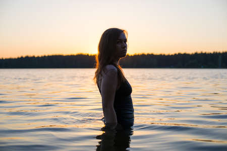 Beautiful Russian girl in a long black swimsuit swims outside the city on the lake in the rays of sunset or dawn. Splashing water on the background of the sun. Happy girl.の写真素材