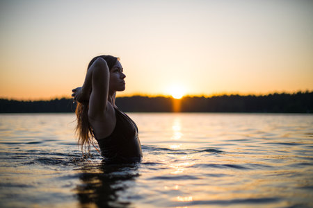 Beautiful Russian girl in a long black swimsuit swims outside the city on the lake in the rays of sunset or dawn. Splashing water on the background of the sun. Happy girl.の写真素材