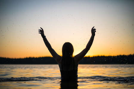 Beautiful Russian girl in a long black swimsuit swims outside the city on the lake in the rays of sunset or dawn. Splashing water on the background of the sun. Happy girl.の写真素材