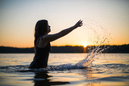 Beautiful Russian girl in a long black swimsuit swims outside the city on the lake in the rays of sunset or dawn. Splashing water on the background of the sun. Happy girl.の写真素材