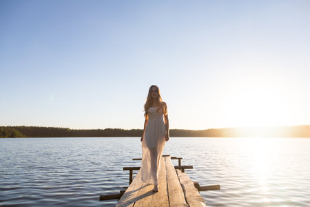 Beautiful Russian girl dressed in a white dress, walking along a wooden pier on the bank of a river or a lakeの写真素材