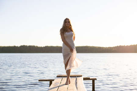 Beautiful Russian girl dressed in a white dress, walking along a wooden pier on the bank of a river or a lakeの写真素材