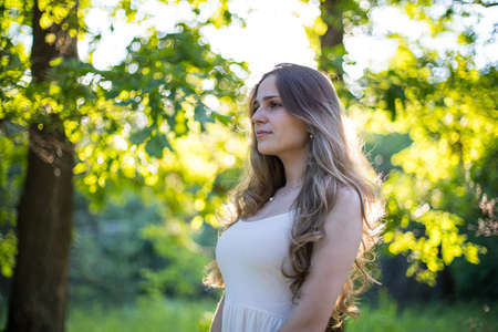Beautiful young Russian girl dressed in a white dress, walks through the forest among the oaks in the rays of the sunsetの写真素材