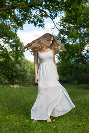 Beautiful young Russian girl dressed in a white dress, walks through the forest among the oaks in the rays of the sunsetの写真素材