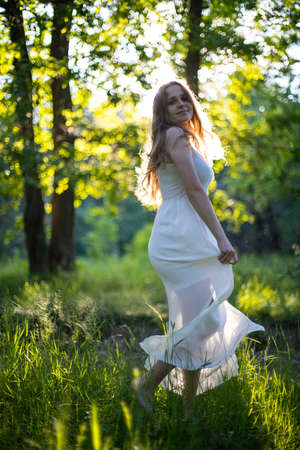 Beautiful young Russian girl dressed in a white dress, walks through the forest among the oaks in the rays of the sunsetの写真素材