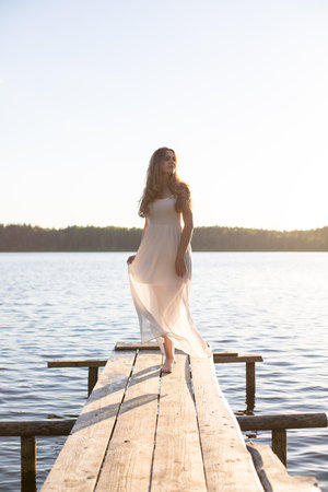 Beautiful Russian girl dressed in a white dress, walking along a wooden pier on the bank of a river or a lakeの写真素材