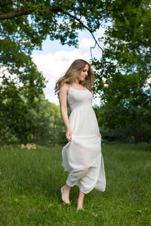 Beautiful young Russian girl dressed in a white dress, walks through the forest among the oaks in the rays of the sunsetの写真素材