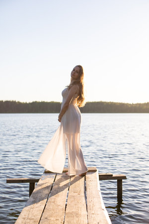 Beautiful Russian girl dressed in a white dress, walking along a wooden pier on the bank of a river or a lakeの写真素材