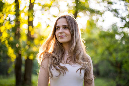 Beautiful young Russian girl dressed in a white dress, walks through the forest among the oaks in the rays of the sunsetの写真素材