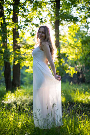 Beautiful young Russian girl dressed in a white dress, walks through the forest among the oaks in the rays of the sunsetの写真素材