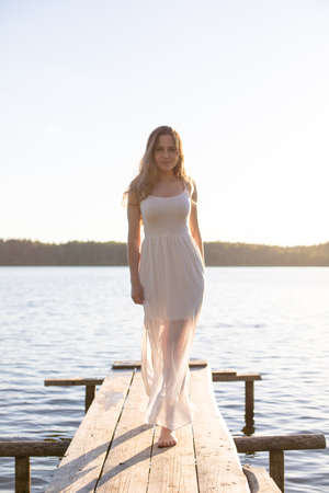 Beautiful Russian girl dressed in a white dress, walking along a wooden pier on the bank of a river or a lakeの写真素材