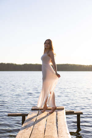 Beautiful Russian girl dressed in a white dress, walking along a wooden pier on the bank of a river or a lakeの写真素材