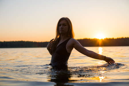 Beautiful Russian girl in a long black swimsuit swims outside the city on the lake in the rays of sunset or dawn. Splashing water on the background of the sun. Happy girl.の写真素材