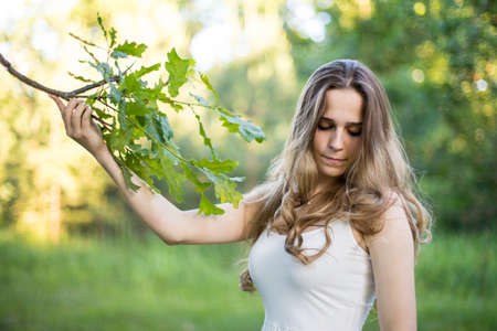 Beautiful young Russian girl dressed in a white dress, walks through the forest among the oaks in the rays of the sunsetの写真素材
