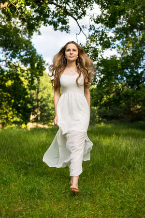 Beautiful young Russian girl dressed in a white dress, walks through the forest among the oaks in the rays of the sunsetの写真素材