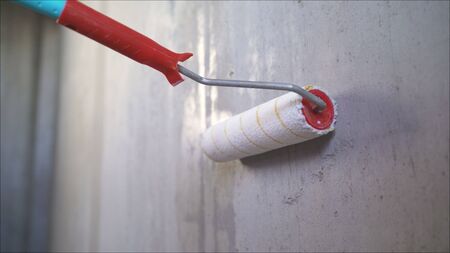 A male hand is applying a primer to a white wall with a roller. Wet concrete wall background. The concept of apartment renovation, construction and finishing work. A worker applies a primer to the wall with a long roller.の写真素材