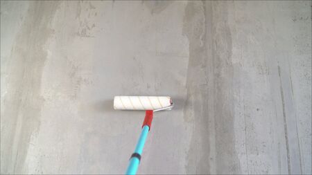 A male hand is applying a primer to a white wall with a roller. Wet concrete wall background. The concept of apartment renovation, construction and finishing work. A worker applies a primer to the wall with a long roller.の写真素材