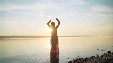 Girl in a light dress on the beach at sunrise. beautiful women in a light pink dress walking along the beach at dawn. Good morning and relaxation. Young beautiful girl stands in the water in a light pink dress on the background of the dawn.の写真素材