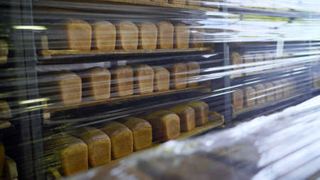 Bread on shelves under cling film. Freshly baked bread trays in a row. The bread is stored in a...