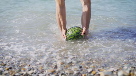 A young guy stands by the sea and holds a ripe watermelon in his hands. The guy gets a watermelon out of the sea.の写真素材