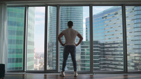 Athletic man looking out the window in the morning. Handsome young guy enjoying beautiful view on a sunny day. Rear back view Close up movement to the window. Athlete on the background of window windows.の写真素材