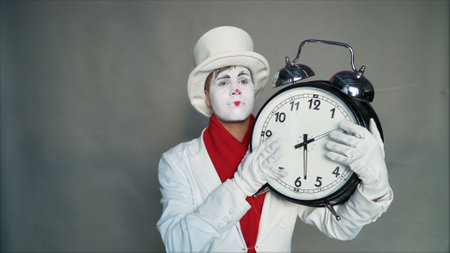 Mime in a suit and hat is holding a big clock. Man in a suit with a dial in his hands. Actor with a painted face and a clock. Time is running out. Time is leaking.の写真素材