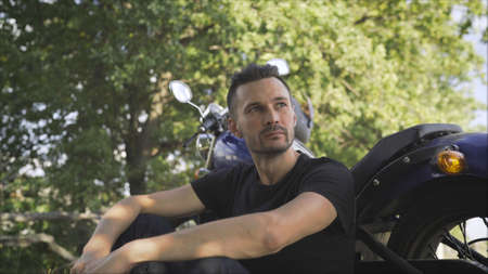 Motorcyclist sits near a motorcycle and looks thoughtfully into the distance.