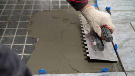 The master applies glue for laying tiles on the concrete floor. A worker levels the adhesive mortar...