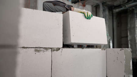 A worker erects a wall of aerated concrete blocks in an apartment. A wall was erected from a modern brick-foam block. The worker places a block on top of another block. We make another layer of the future wall.の写真素材