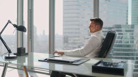 Pensive man sitting in a chair at the table. Expensive study on a high floor in a skyscraper. A businessman in a white shirt is sitting in his expensive office on a high floor.の写真素材