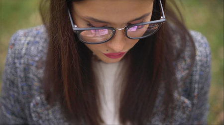 Close-up view of a girl with glasses reading a book. Look at the glasses below. A girl in a coat looks in a book, close-up. Outdoors lifestyle fashion portraitの写真素材