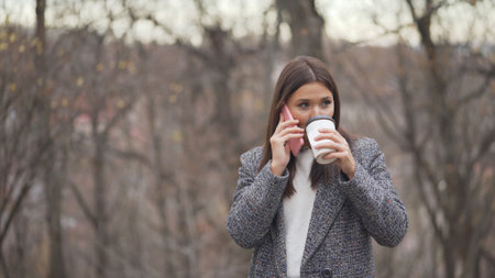 Young girl outdoors in a coat talking on the phone and drinking coffee. Beautiful young woman talking on mobile phone while drinking coffee outdoorsの写真素材