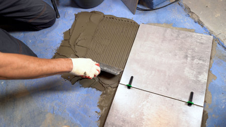 Tile adhesive for bonding ceramic tiles to the floor. Applying tile adhesive to the floor when laying floor tiles by a tiler. Bathroom renovation with tile adhesive. A worker lays dark gray ceramic floor tiles on an adhesive surface.の写真素材