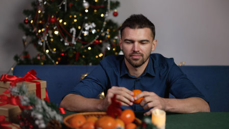 A guy at the New Years table, against the backdrop of a decorated Christmas tree. A guy at the festive table peels a tangerine. New Year celebration concept.の写真素材