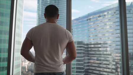 Athlete on the background of window windows. Athletic man looking out the window in the morning. Handsome young guy enjoying beautiful view on a sunny day. Rear back view Close up movement to the windowの写真素材