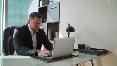 Guy in a suit at a laptop in his office. A tense businessman is sitting at a laptop against the...