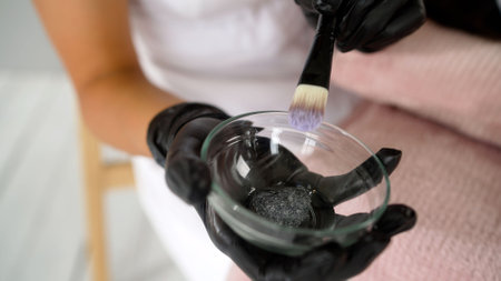 Beautician wearing black gloves mixes a charcoal face mask in a glass bowl, preparing for a rejuvenating skincare treatment.の写真素材