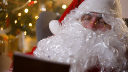 Santa Claus writes a letter on paper while sitting on an armchair near the Christmas tree. Santa...