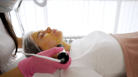 Beautician applying cosmetic mask on a client face lying on a cosmetology bed.