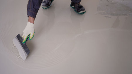 A worker spreads liquid on the floor with a spatula. Leveling liquid self-leveling floor screed. A worker spreads liquid cement mortar on a construction site. Leveling a liquid floor with a spatula.の写真素材