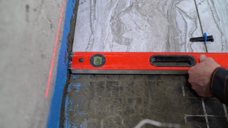 Construction worker is using a spirit level while laying tile on a floor, ensuring that it is level.