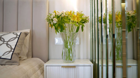 Fresh bouquet of yellow and white flowers sits in a glass vase on a white nightstand in a modern and...