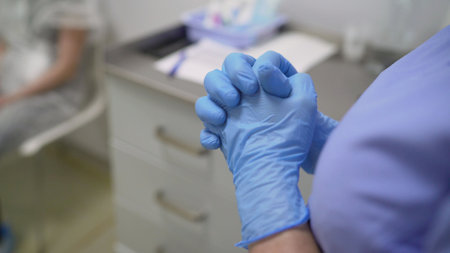 Doctor clasping hands while wearing blue surgical gloves with patient sitting in background in medical officeの写真素材