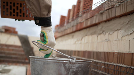 Construction worker holding a brick in one hand and picking up a trowel from a bucket of mortar while building a brick wallの写真素材