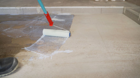 Skilled construction professional smoothing epoxy resin coating on concrete floor using specialized paint roller, applying protective industrial surface treatmentの写真素材