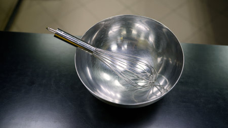 Metal whisk resting in stainless steel mixing bowl on black countertop, ready for cooking and bakingの写真素材