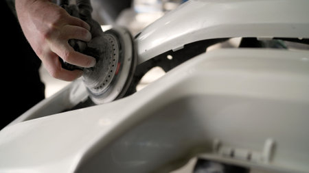 Car mechanic skillfully polishing a white car bumper with an orbital sander in a busy professional workshop, focusing on precision and detailの写真素材
